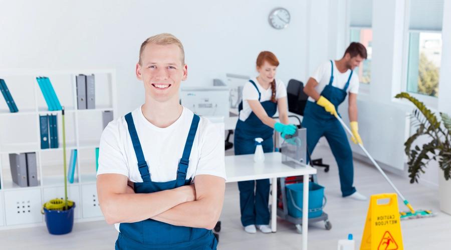 Smiling cleaner in blue overalls in an office, with two colleagues cleaning in the background